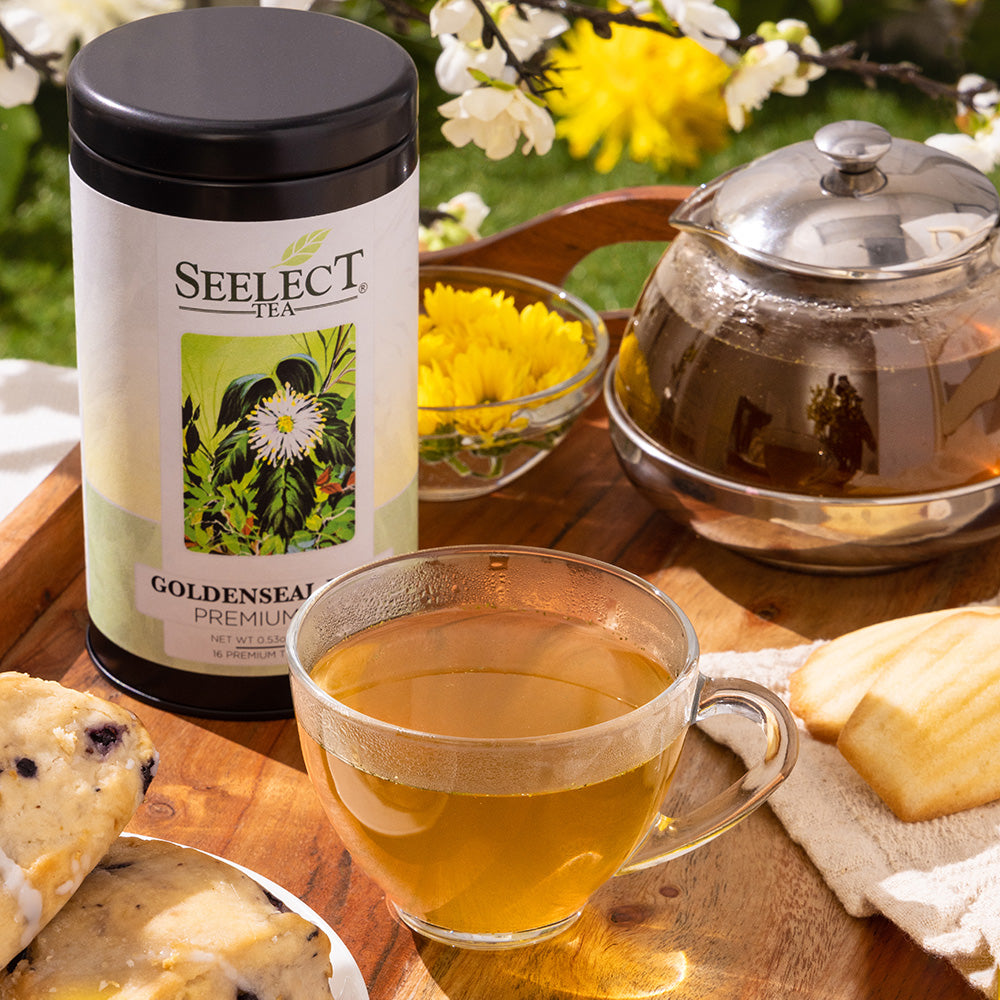 Tea set with a cup of tea, teapot, and 'Seelect Tea' Goldenseal tea container on a wooden table with flowers in the background.