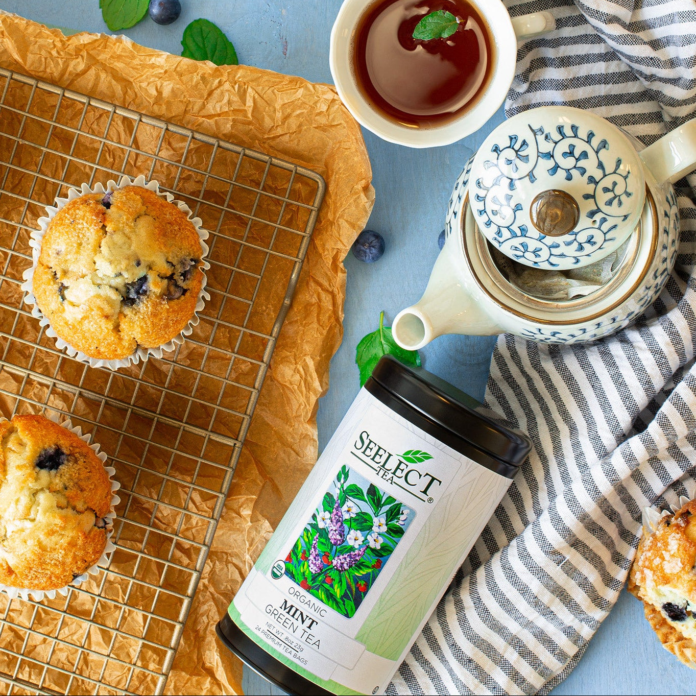 Blueberry muffins on a cooling rack with tea, mint leaves, and a bottle of mint tea on a blue surface.