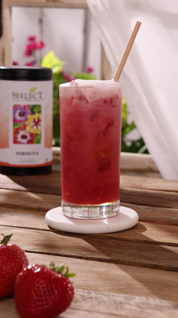 Pink iced drink with a straw in a glass on a wooden surface, surrounded by strawberries and a bottle of Select tea.