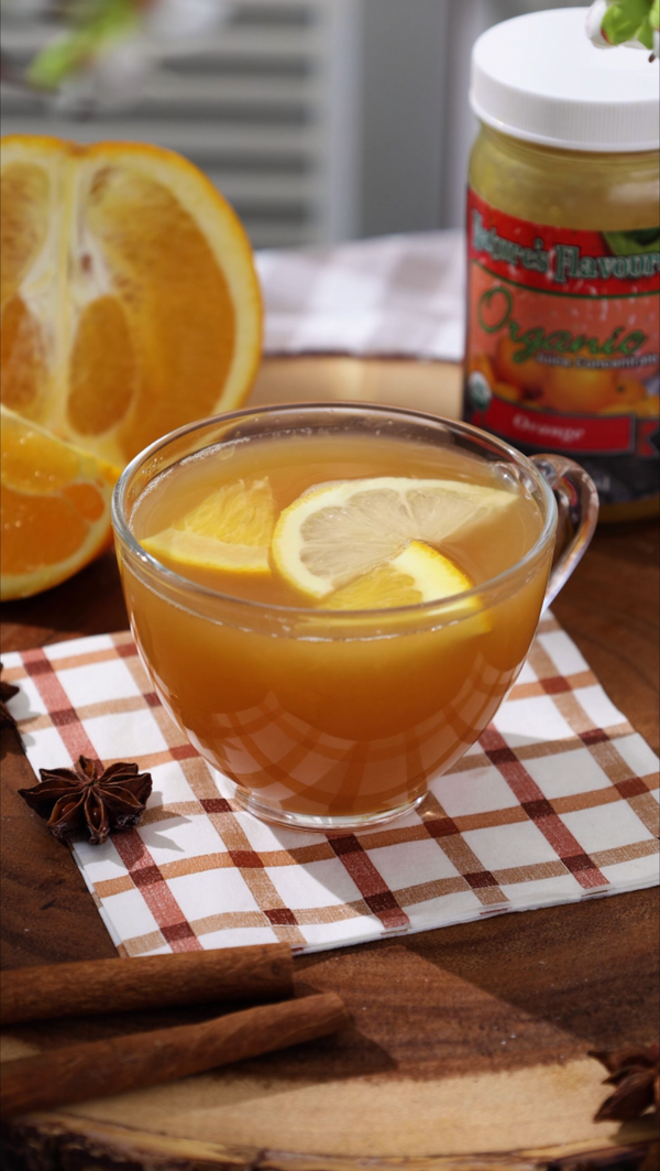 Glass of tea with citrus slices on a checkered cloth, with oranges and a jar in the background.