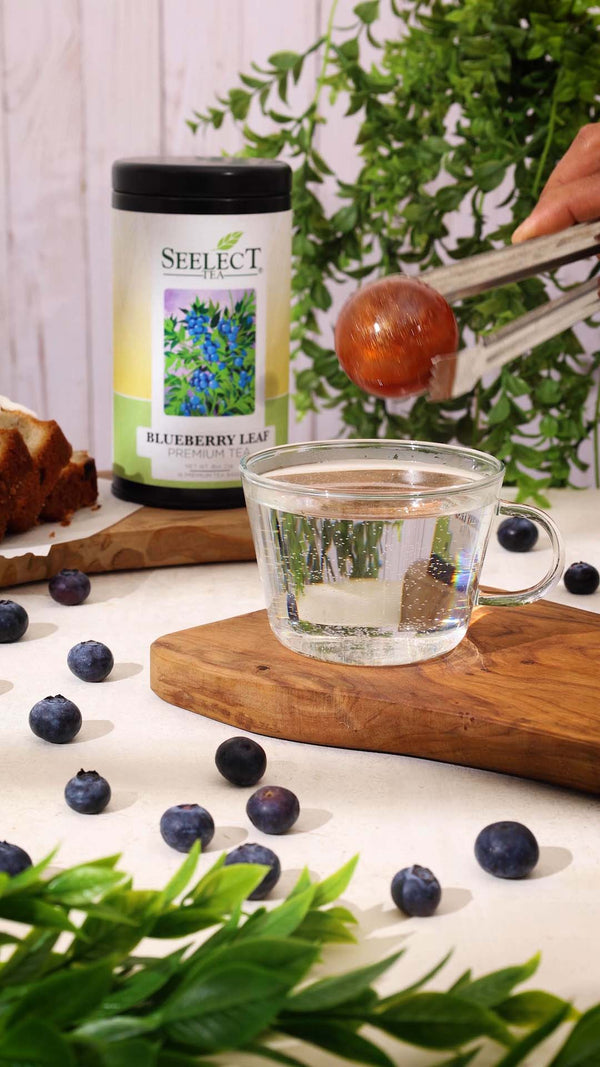 Person putting tea into a glass of water next to a jar labeled 'Select' with blueberries and green leaves on a white surface.